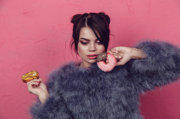 Beautiful girl in a fur coat with donuts in hands posing on a pink background. - Foto, imagen
