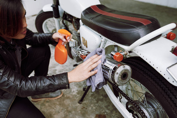 Συμπυκνωμένο φορώντας δερμάτινο μπουφάν ποδηλάτης χρησιμοποιώντας κουρέλι και καθαρισμό ψεκασμού για να σκουπίσετε τη βρωμιά από μοτοσικλέτα του - Φωτογραφία, εικόνα