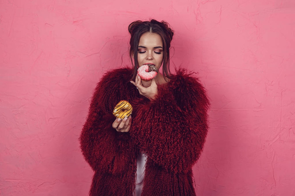 Girl in a bright fur coat with donuts in hands posing on a pink background. - Foto, afbeelding