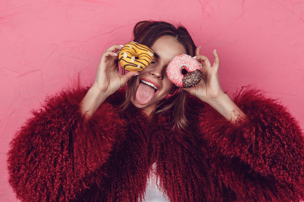 Girl in a bright fur coat with donuts in hands posing on a pink background. - Valokuva, kuva
