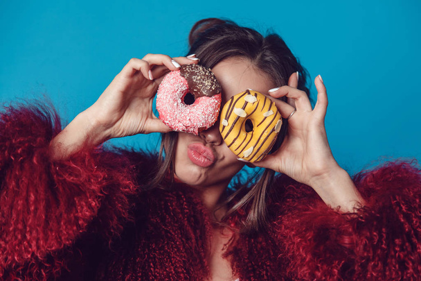 Attractive young woman in bright fur coat posing with donuts in hands. - Фото, изображение