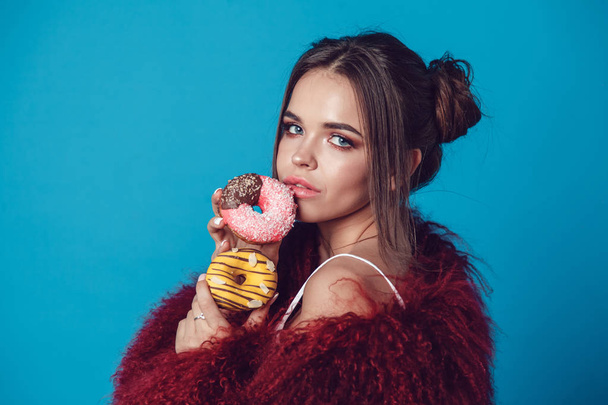 Attractive young woman in bright fur coat posing with donuts in hands. - Photo, Image