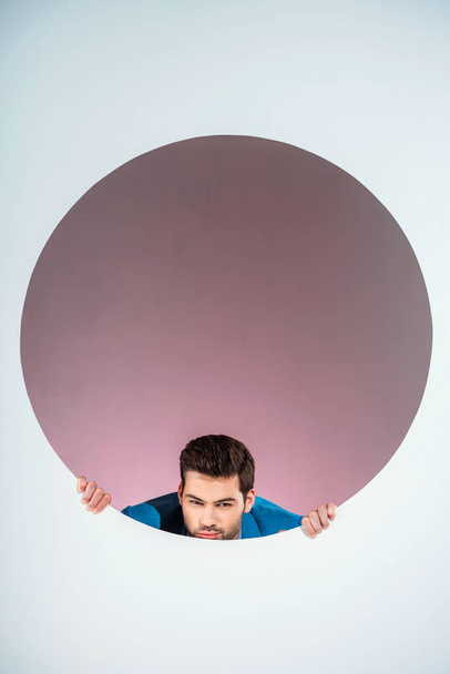stylish bearded young man looking at camera through hole on grey  - Foto, immagini