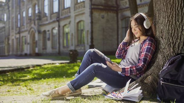 Multiracial girl sitting under tree in headphones, scrolling screen of cellphone - Photo, image