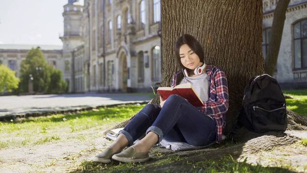 Mixed female sitting under tree, reading favorite book, gripping plot, engrossed - Foto, imagen