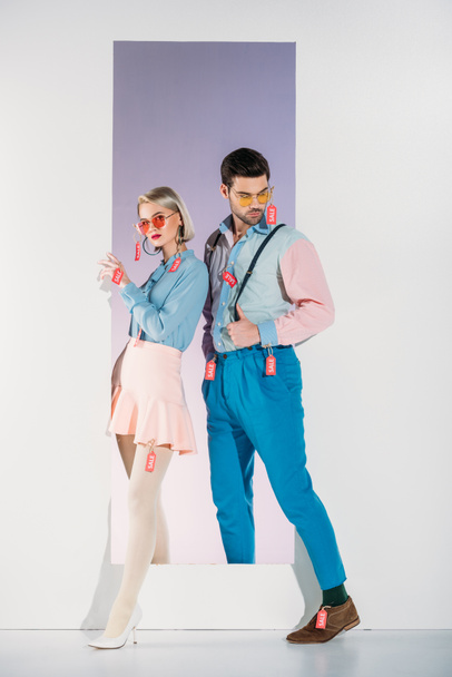 full length view of stylish young couple with sale tags on clothes standing in opening on grey  - Photo, Image