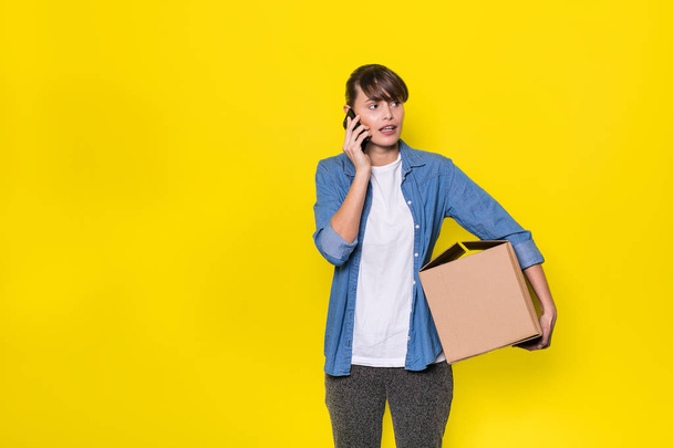 pretty woman standing on yellow background with moving cardboard box and using her cellphone - Zdjęcie, obraz