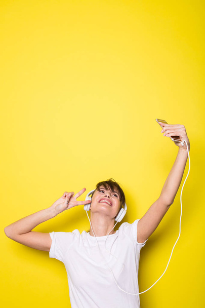 jonge en mooie vrouw luisteren muziek met witte koptelefoon op geïsoleerde op gele achtergrond - Foto, afbeelding