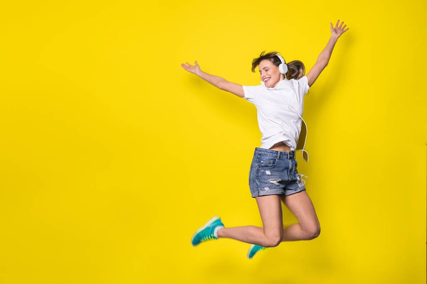 young and happy woman listening music and jumping with big smile on yellow background - Foto, Imagen
