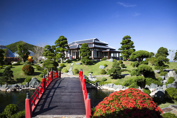 川と周辺の緑の植物がたくさんある日本庭園の伝統的な日本スタイルの美しい建物にかかる赤いレトロな橋 - 写真・画像