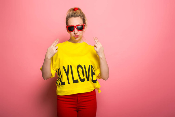 Jovem mulher expressiva vestindo camiseta amarela brilhante e óculos de sol no fundo rosa
 - Foto, Imagem
