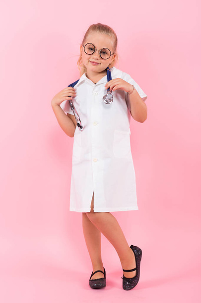 Cute little girl in medical uniform with stethoscope on pink background - 写真・画像