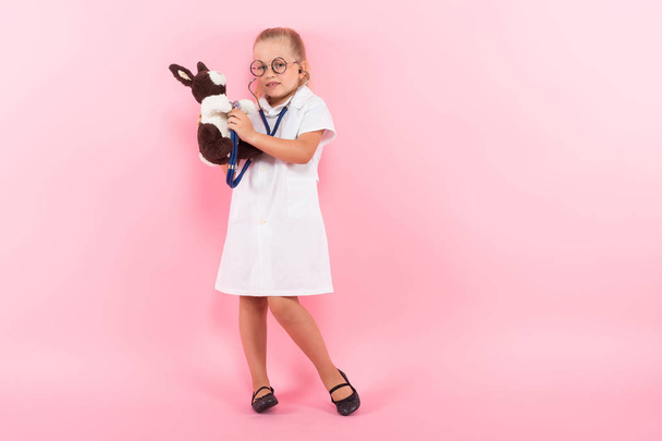 Cute little girl in medical uniform with stethoscope and plush rabbit on pink background - Фото, зображення