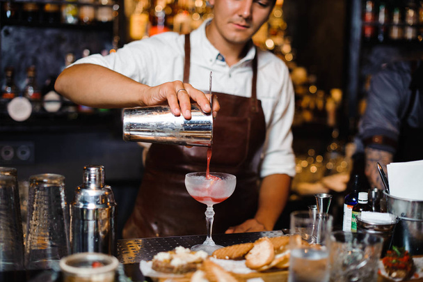 Barman in a brown leather apron pouring fruit alcoholic cocktail into the glass standing behind the bar counter - Foto, Imagen