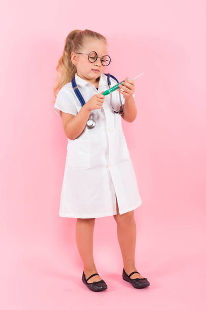 Cute little girl in medical uniform with stethoscope and syringe on pink background - Photo, Image