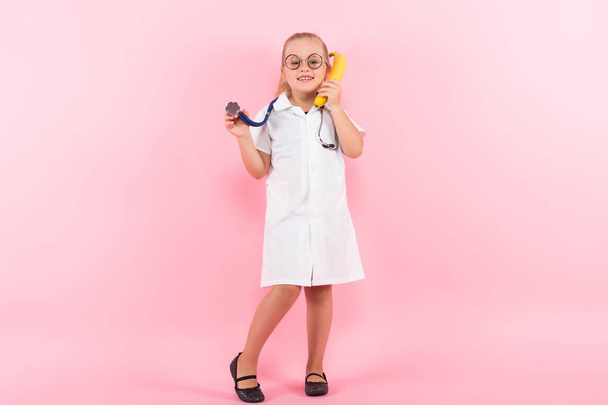 Cute little girl in medical uniform with stethoscope and banana on pink background - Foto, Imagem
