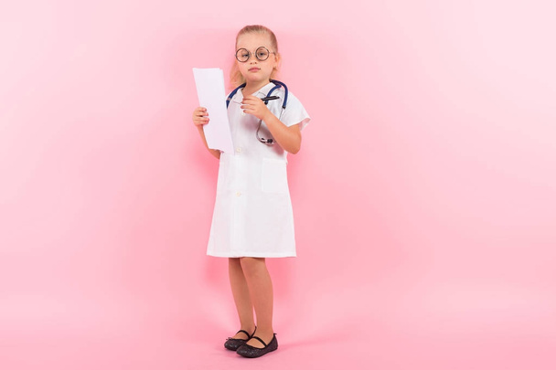 Cute little girl in medical uniform with stethoscope, white sheets and pen on pink background - Foto, Imagem