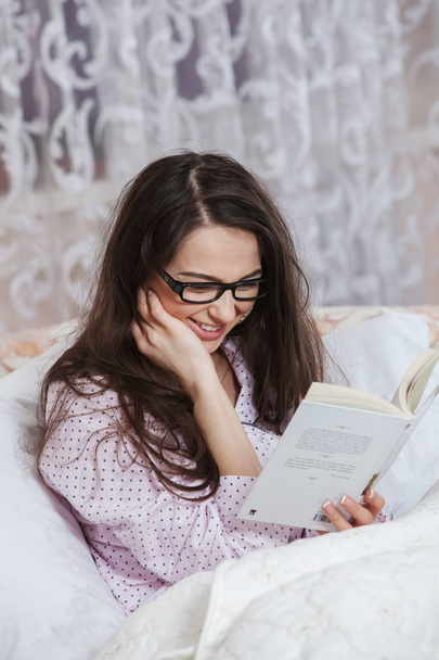 Młoda kobieta, noszenie okularów podczas czytania książki w łóżku. Zrelaksowany kobietę leżącą w łóżku w piżamy, czytanie książki. - Zdjęcie, obraz
