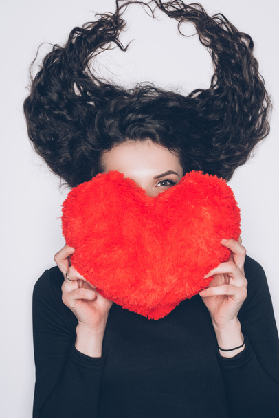 young woman with hair in shape of horns covering face with heart shaped pillow isolated on white - Foto, Bild
