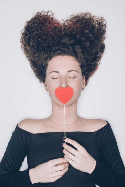 attractive young woman with curly hair covering mouth with red heart isolated on white, st valentines day concept - Fotó, kép