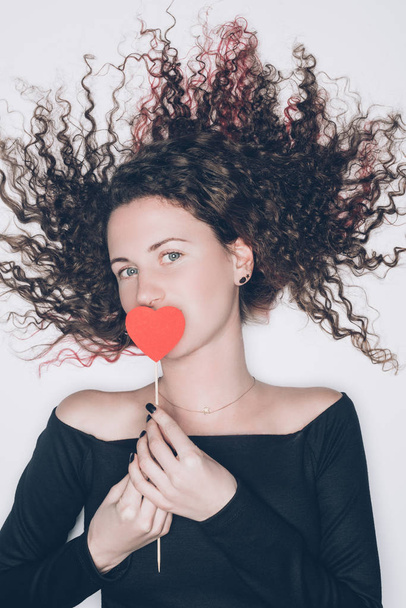young woman with curly hair covering mouth with red heart isolated on white - Foto, Imagem
