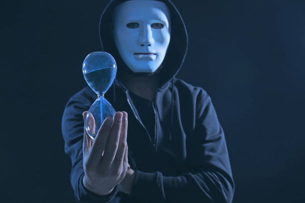 Masked hacker with sand clock in darkness - 写真・画像