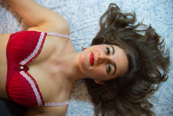 Piękna Brunetka kobieta stwarzających w czerwony biustonosz. Leżąc na poduszce, czerwone usta, brązowe włosy. Czerwony biustonosz z białe brzegi.  - Zdjęcie, obraz