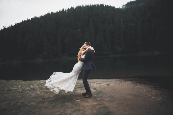 Young newly wed couple, bride and groom kissing, hugging on perfect view of mountains, blue sky - Фото, изображение