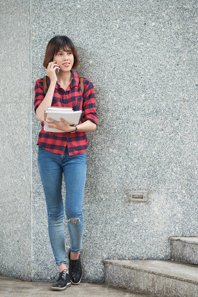 Azjatki, młoda kobieta z książek w jej ręce, opierając się na ścianie i rozmawia przez telefon - Zdjęcie, obraz