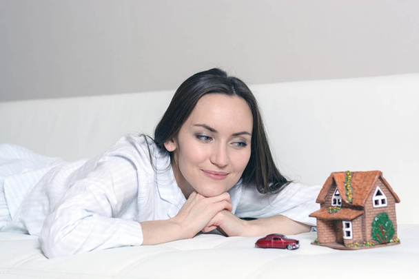Νεαρή γυναίκα στο πιτζάμες που βρίσκεται σε ένα λευκό καναπέ σε ένα δωμάτιο μπροστά από Wendy ένα σπίτι και ένα αυτοκίνητο, και ονειρεύεται ένα λαμπρό μέλλον. - Φωτογραφία, εικόνα
