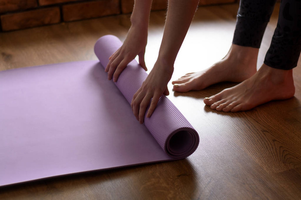 Uma mulher descalça torce um tapete de ioga roxo e fitness no chão do parquete - Foto, Imagem