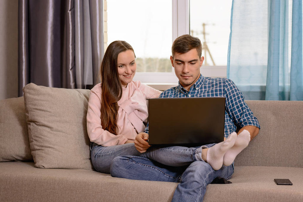 A young couple sitting on the couch in the room and watch the video on the laptop on his knees - Foto, imagen