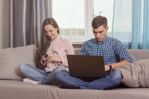 Young couple sitting on the sofa in the room, busy with their gadgets, communication problems of modern society - Foto, Imagen