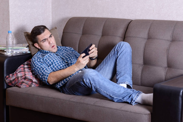 Bild erregt von einem borstigen Mann in Hemd mit Käfigprint, Jeans und weißen Socken, liegt zu Hause auf dem Sofa und spielt am Smatphone. Leidenschaft für das Spiel - Foto, Bild