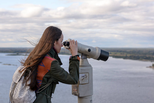 A young woman traveler on the observation deck looking through binoculars at the panorama of the city of Nizhny Novgorod - Photo, Image