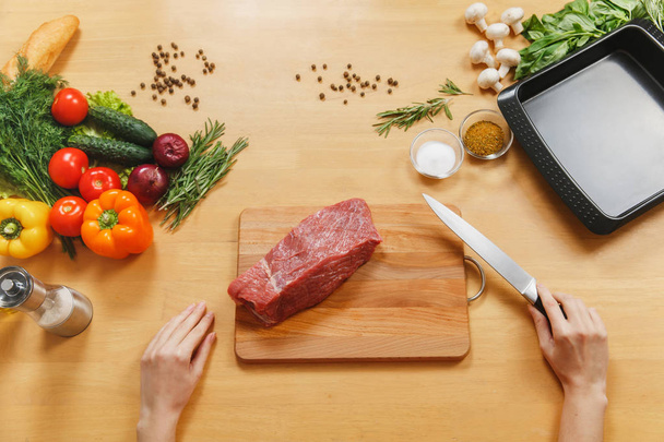 Raw meat. Fresh beef tenderloin on cutting board on wooden table with different vegetables, spices, mushrooms, baking tray, knife. Top view flatlay. Copy space for advertisement. Woman cuts with knife - Photo, Image