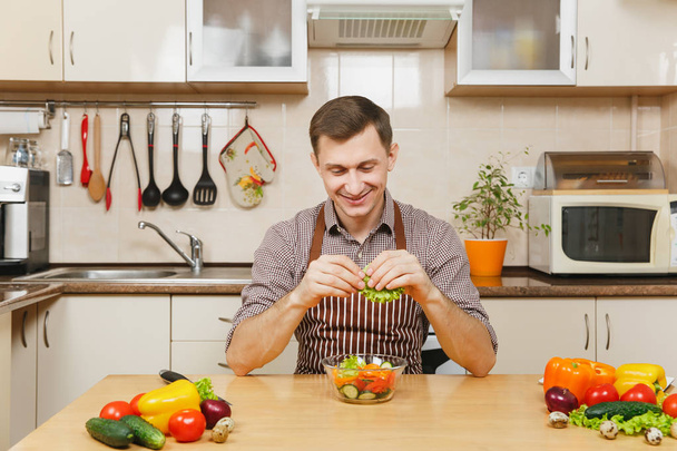 Kavkazské pohledný mladý muž v zástěře, hnědá košile sedí u stolu, síla a trhání salát zelenina na salát s nožem v lehké kuchyni. Pojem dieta. Vaření doma. Příprava potravin. - Fotografie, Obrázek