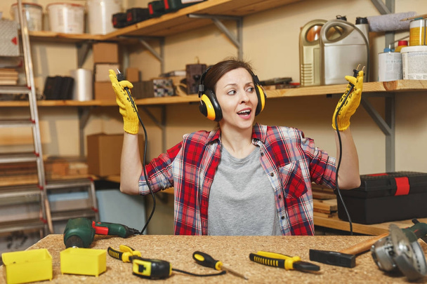 Beautiful crazy fun caucasian young woman in plaid shirt, noise insulated headphones working in carpentry workshop at table place, with two wires from electrical appliances like power drill and saw. - Photo, Image