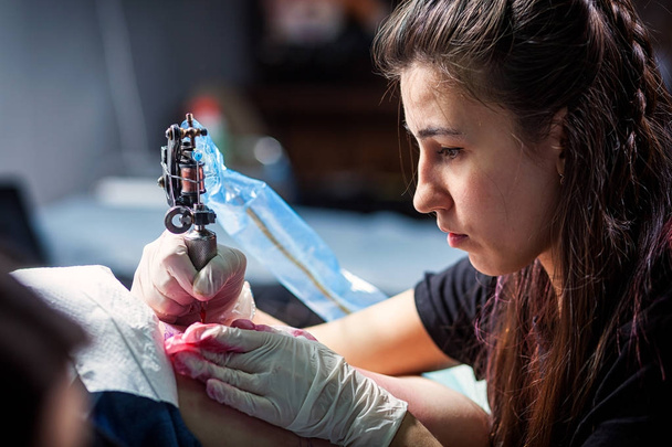 Joven mujer morena tatuaje maestro haciendo tatuaje de un pequeño pájaro con máquina de tatuaje rojo, primer plano
 - Foto, Imagen