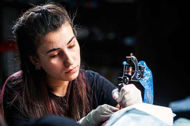 Közeli női professzionális tetováló mester csinál egy kis madárka tetoválás mell piros festékek vonzó nő a sötét tattoo szalonban, oldalnézetből - Fotó, kép