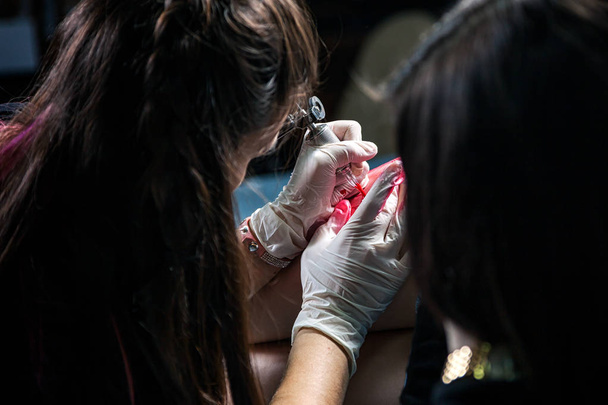 Ένα κοντινό πλάνο της διαδικασίας δημιουργώντας ένα τατουάζ της ένα πουλί με κόκκινες χρωστικές ουσίες, χρησιμοποιώντας ένα μηχάνημα τατουάζ από τον πλοίαρχο γυναικεία τατουάζ στο ένα ισχίο υπό στείρες συνθήκες - Φωτογραφία, εικόνα