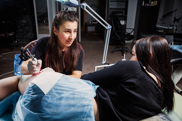 Una giovane tatuatrice che fa un tatuaggio con una macchina per tatuaggi sotto la luce brillante di una lampada su una bella donna con cui parla in un laboratorio buio
 - Foto, immagini