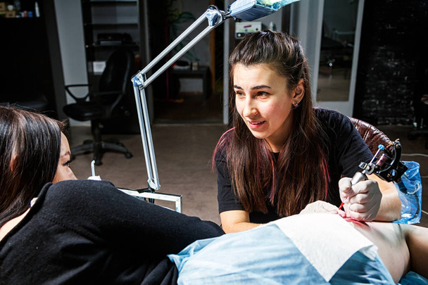 Una joven artista del tatuaje haciendo un tatuaje con una máquina de tatuaje bajo la luz brillante de una lámpara en una hermosa mujer con la que habla en un taller oscuro
 - Foto, Imagen