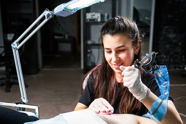 Nuori nainen tatuoija hymyilee ja tekee tatuointia tatuointikoneella lampun kirkkaassa valossa pimeässä työpajassa
 - Valokuva, kuva
