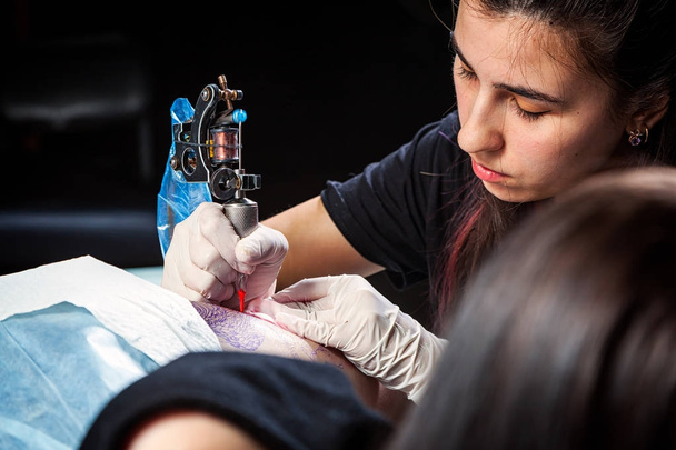 Master tatuointi piirtää punaista maalia asiakkaiden tatuointiin. Tatuoija, jolla on metallinen tatuointikone steriileissä käsineissä ja työskentelee ammattimaisella sinisellä matolla
. - Valokuva, kuva