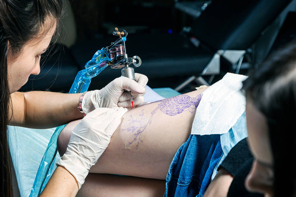 Προσωπογραφία γυναίκας τατουάζ πλοίαρχος δείχνοντας μια διαδικασία δημιουργίας τατουάζ σχετικά με το μηριαίο οστό κάτω από το φως λαμπτήρων. - Φωτογραφία, εικόνα