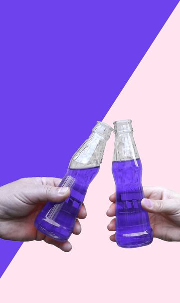 Dos manos sosteniendo una bebida y haciendo tostadas, botellas tintineo con limonada en el fondo del color de moda del año ultra violeta 18-3838 TCX y blanco
 - Foto, Imagen