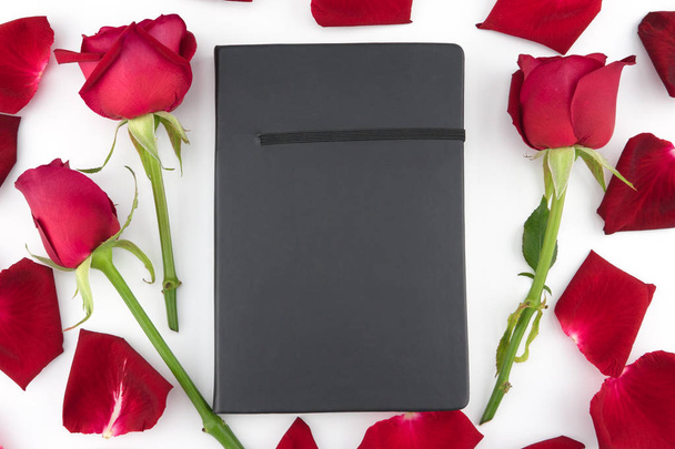 Caderno preto decorado com rosas vermelhas e pétalas sobre fundo branco
 - Foto, Imagem