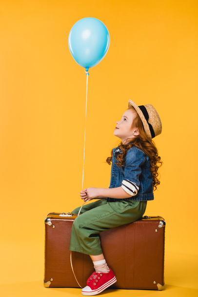 side view of kid with balloon sitting in suitcase isolated on yellow - Zdjęcie, obraz