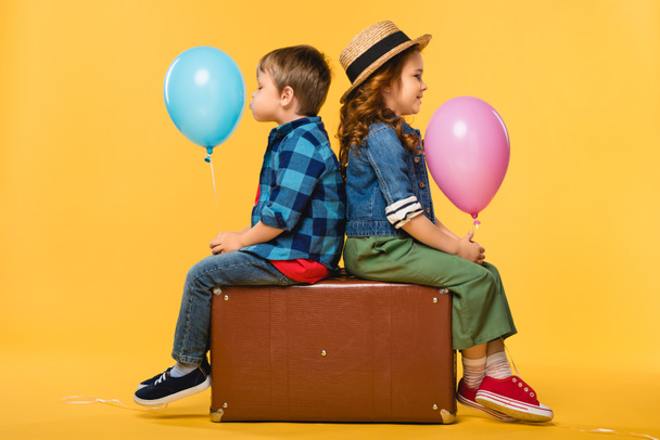 πλαϊνή θέα των παιδιών με μπαλόνια που κάθεται σε δερμάτινη βαλίτσα που απομονώνονται σε κίτρινο - Φωτογραφία, εικόνα